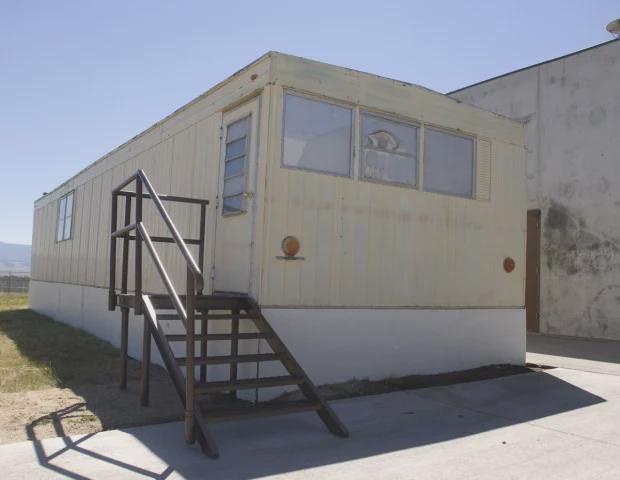 A beige mobile home with a metal staircase leading to its entrance, set against a clear sky. A cat is visible looking out of…