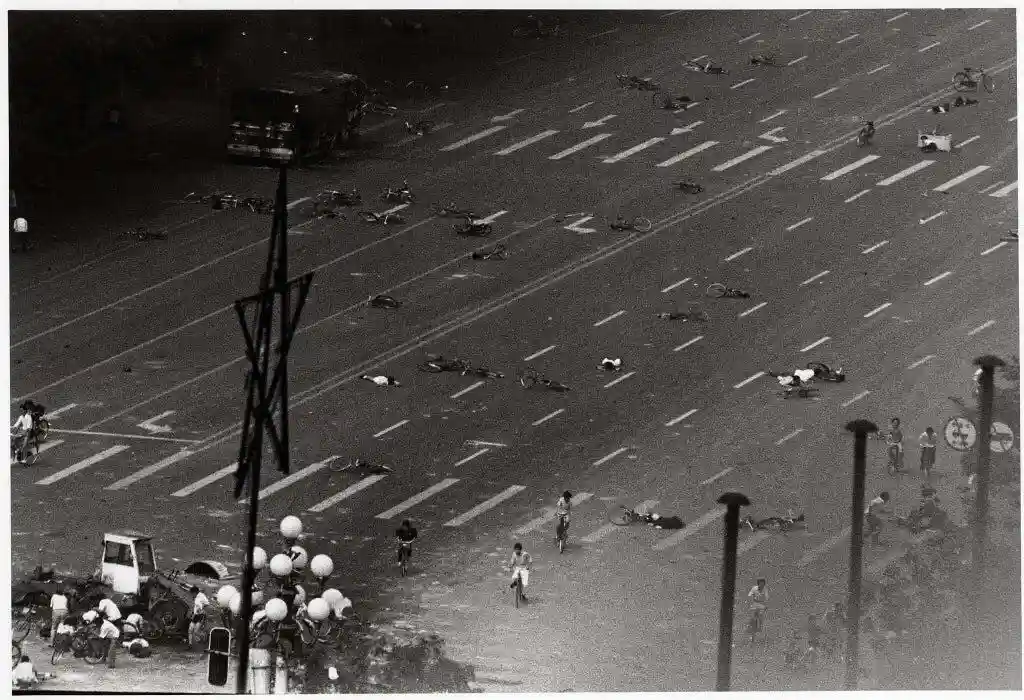 a black and white photograph shows a wide street with numerous people and bicycles scattered across the road, many lying on…