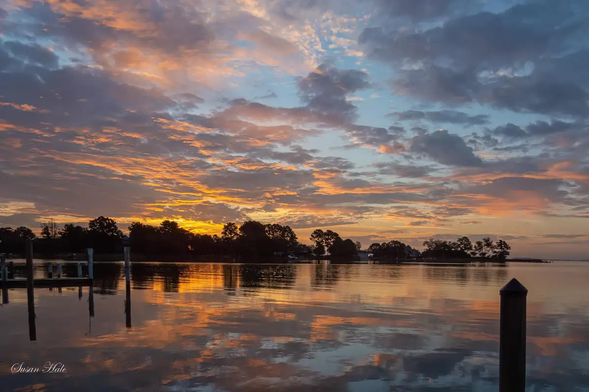 A serene sunset over a calm body of water, with vibrant orange and pink clouds reflected in the surface. Silhouetted trees…
