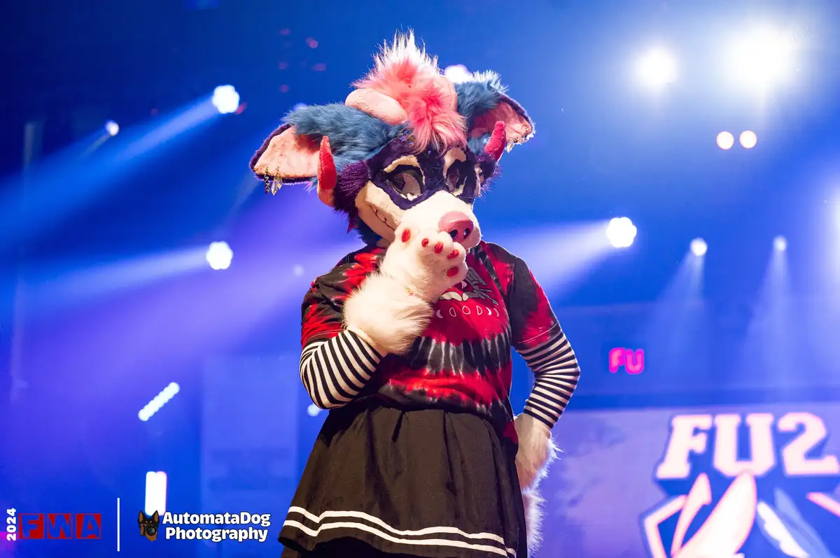 A person in a colorful fursuit with blue and pink hair, red horns, and a pig-like snout stands on a stage under blue lights…