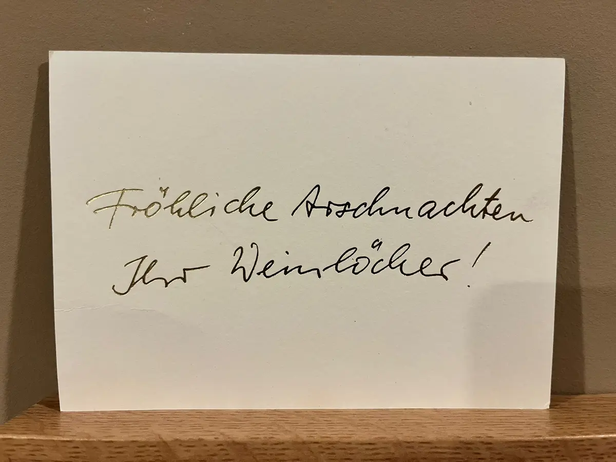 A white greeting card with cursive handwriting in German sits on a wooden surface against a brown wall. The text reads…