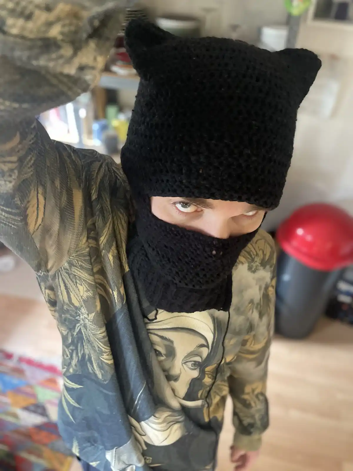 A person wearing a black knitted cat-ear beanie that covers their face except for their eyes, looking intensely at the…