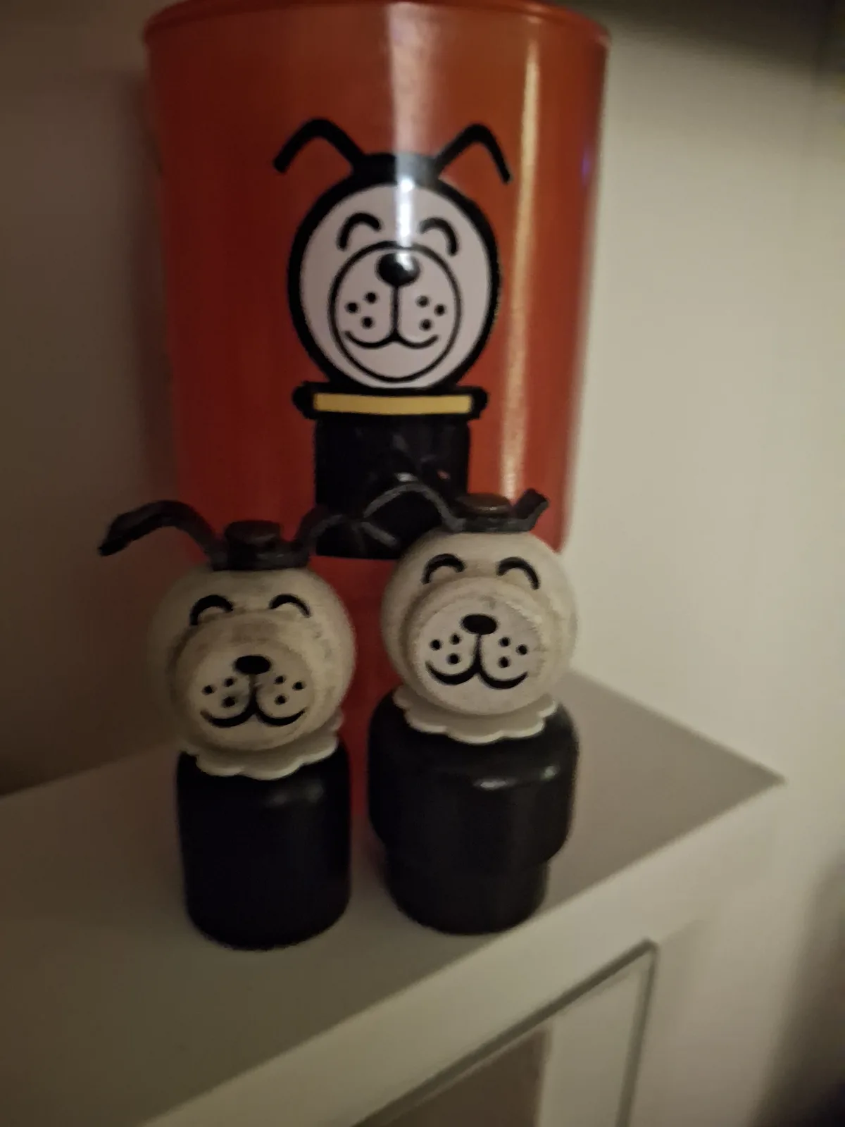 Three dog-themed objects are arranged on a shelf: a large orange cup with a cartoon dog face and a smaller black dog figure…