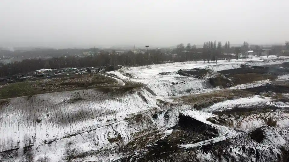 An aerial view of a snowy, barren, excavated landscape with visible tire tracks and distant buildings under a gray sky…