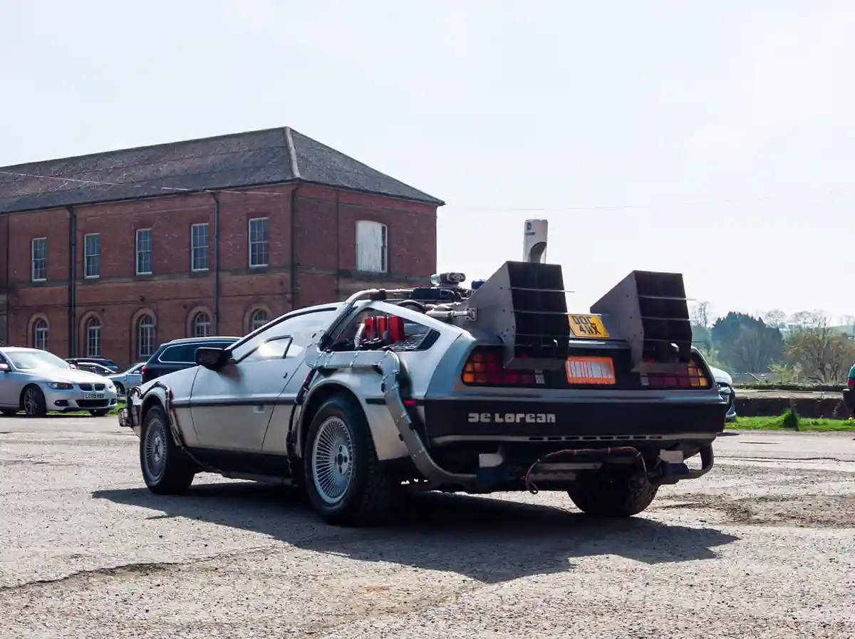 A silver DeLorean car with modified rear wings and exposed mechanical parts is parked in a gravel lot in front of a red…