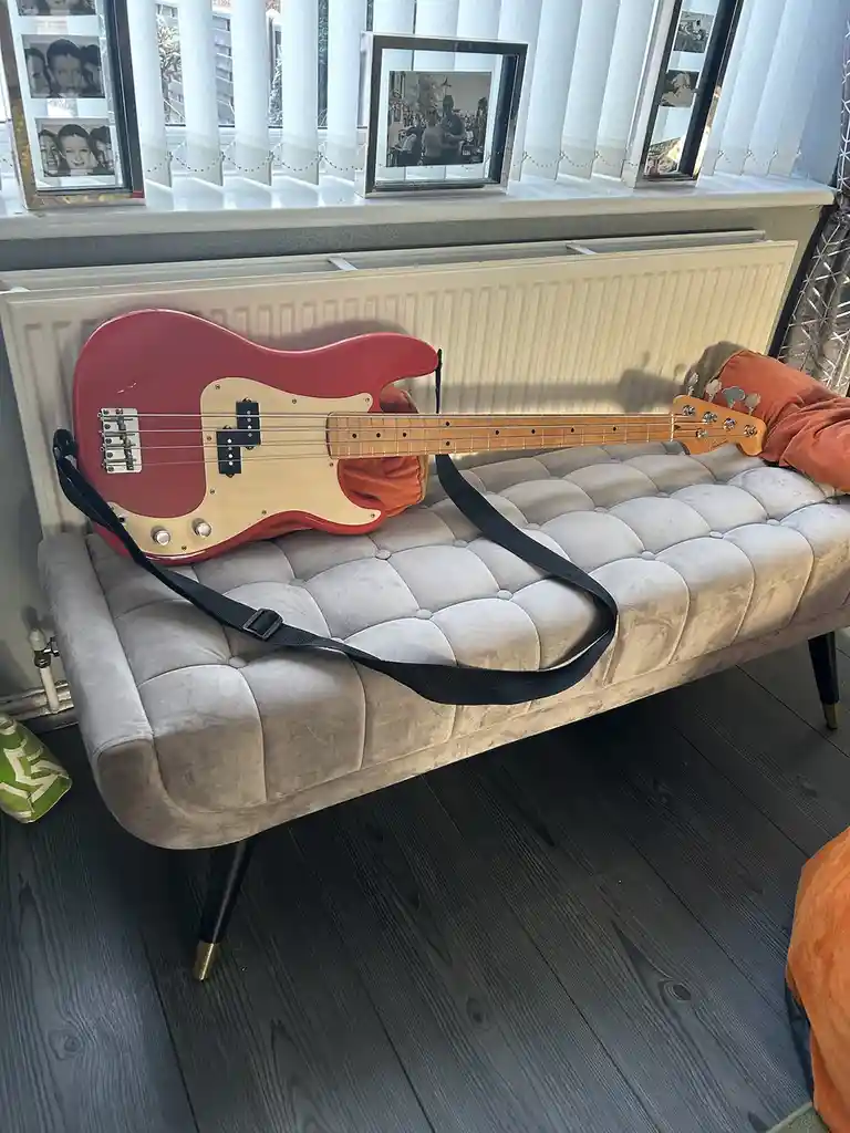 A red and natural wood finish bass guitar with a black strap is resting horizontally across a light gray tufted velvet…