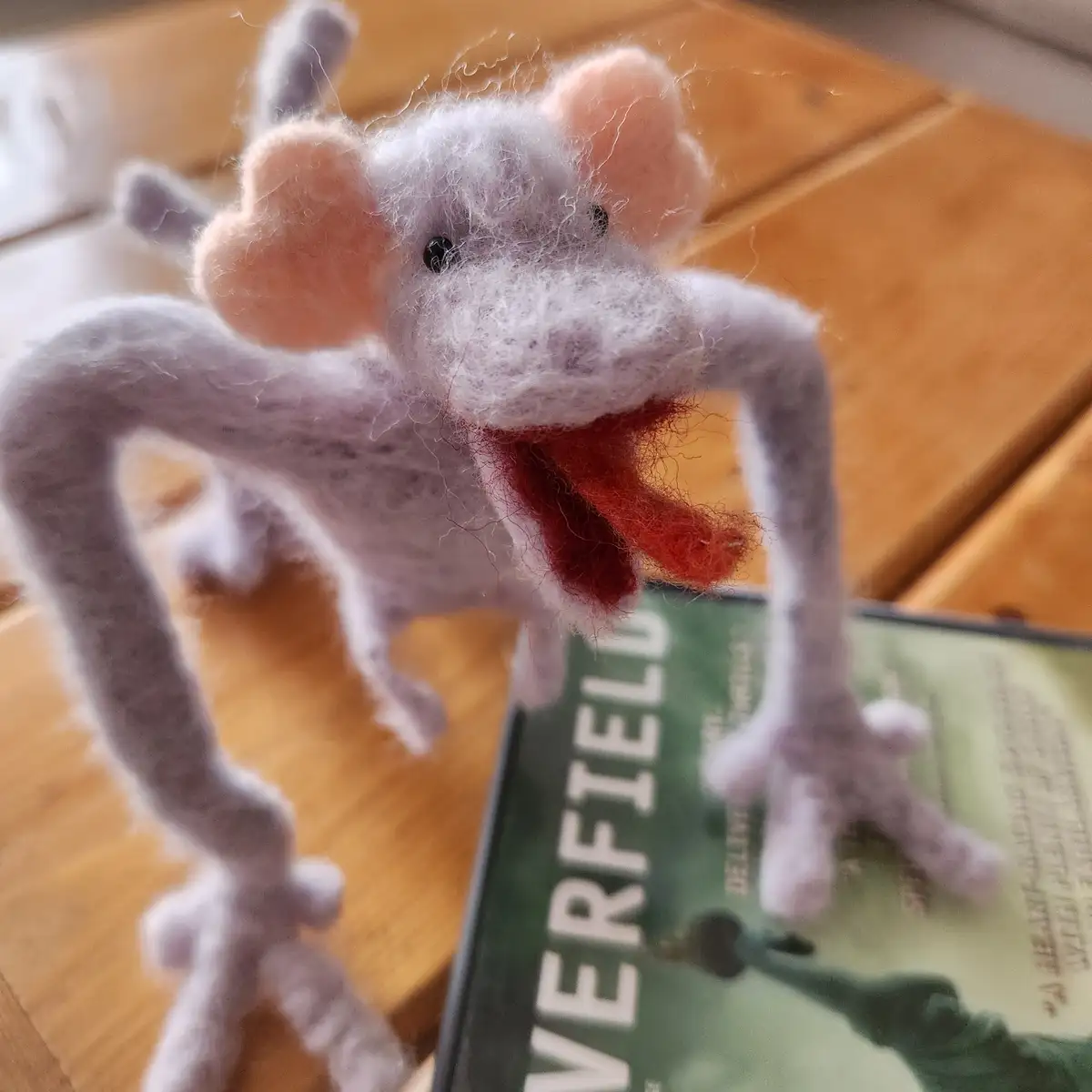 A handmade, fuzzy, purple felt creature with long limbs and a red tongue sticking out stands on a wooden surface next to a…