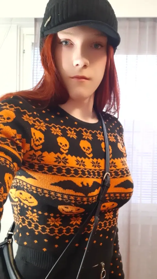 A person with red hair and a black cap wears a black and orange sweater with Halloween motifs like skulls, bats, and…