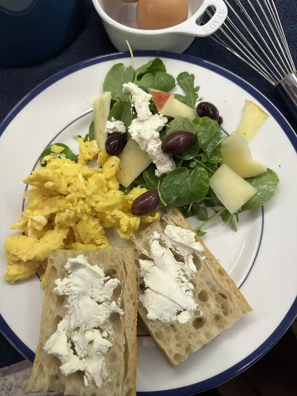 A plate of breakfast food featuring scrambled eggs, a green salad with apple slices, olives, and crumbled white cheese, and…