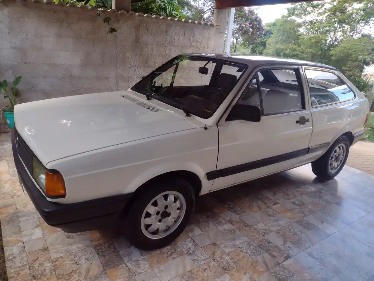 A white 1984 Volkswagen Gol hatchback is parked on a tiled patio under a covered area, with a textured wall and greenery in…