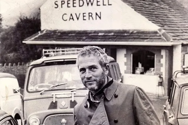A black-and-white photo shows a man with a beard and short hair standing in front of a building with a sign that reads…