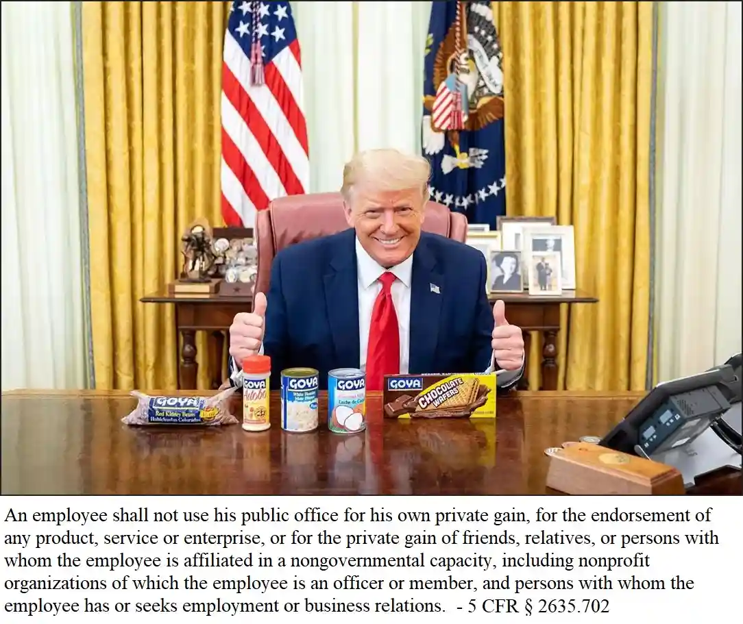 A man in a suit and red tie sits at a desk in an office with American flags and curtains behind him, giving two thumbs up…