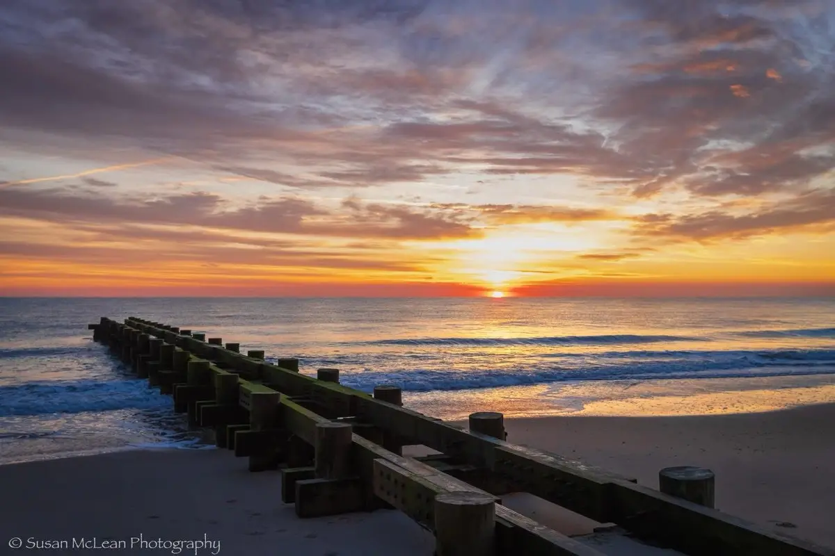 A serene sunset photograph shows a wooden breakwater extending into the ocean, with vibrant orange, yellow, and purple…