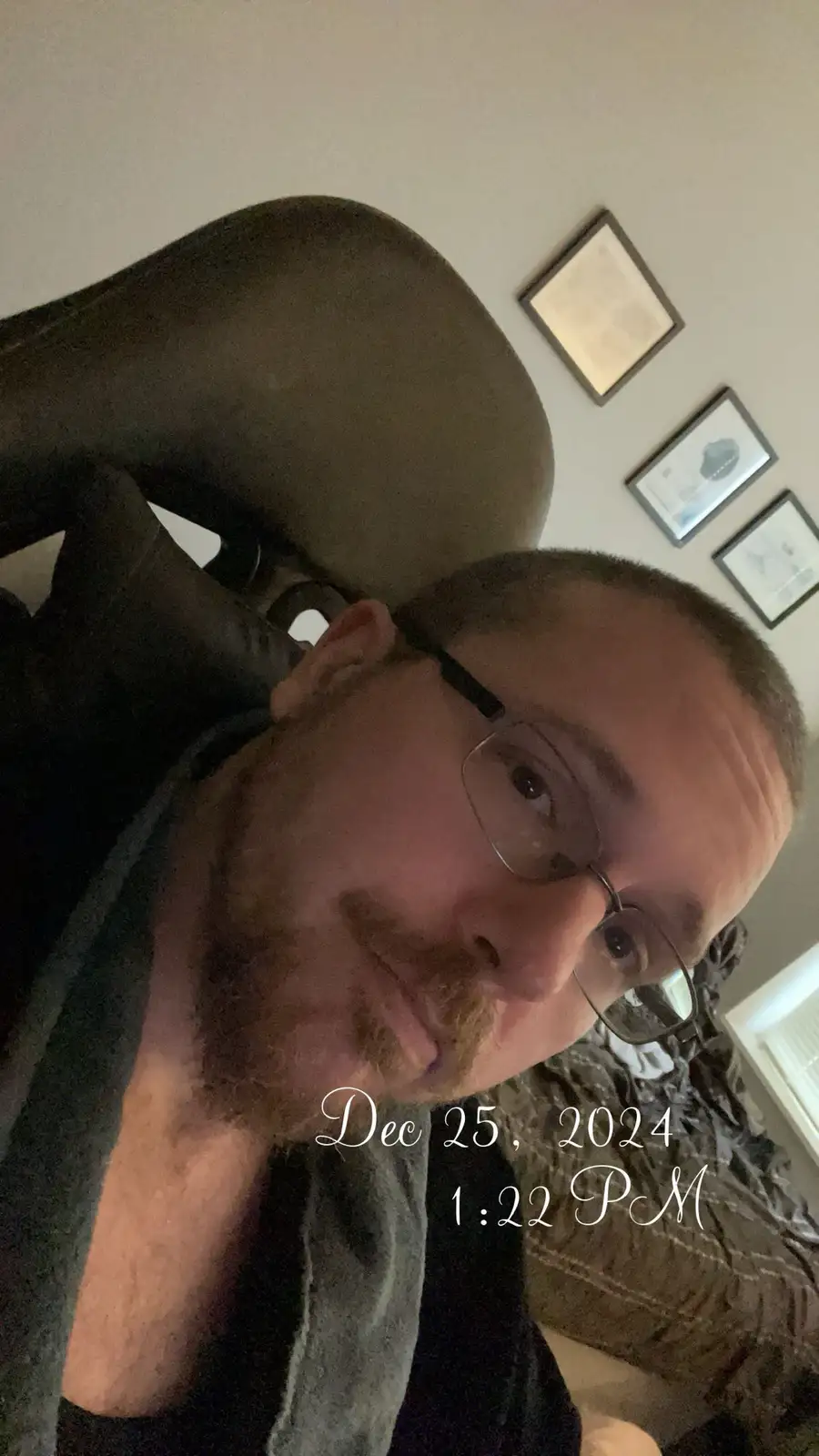 A man with glasses and a short beard is taking a selfie while reclined in a dark chair, looking directly at the camera with…