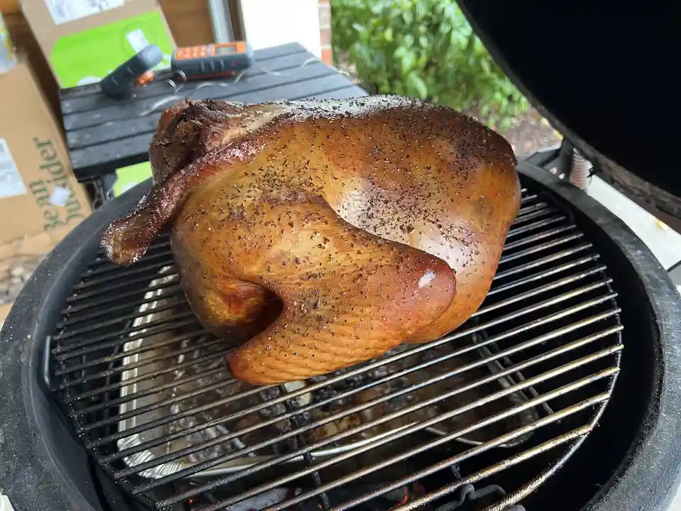 A whole roasted bird, likely a turkey or chicken, sits on the grates of a charcoal grill with a golden-brown, seasoned skin…