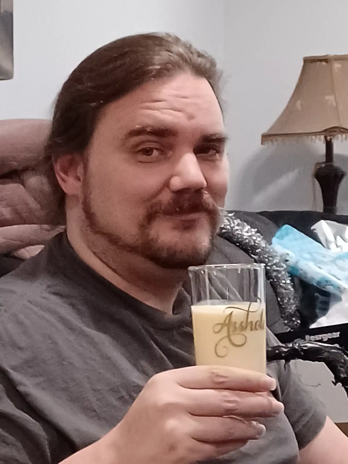 A man with long dark hair and a mustache sits on a couch holding a glass of a creamy beverage, which has the word 'Asshole'…