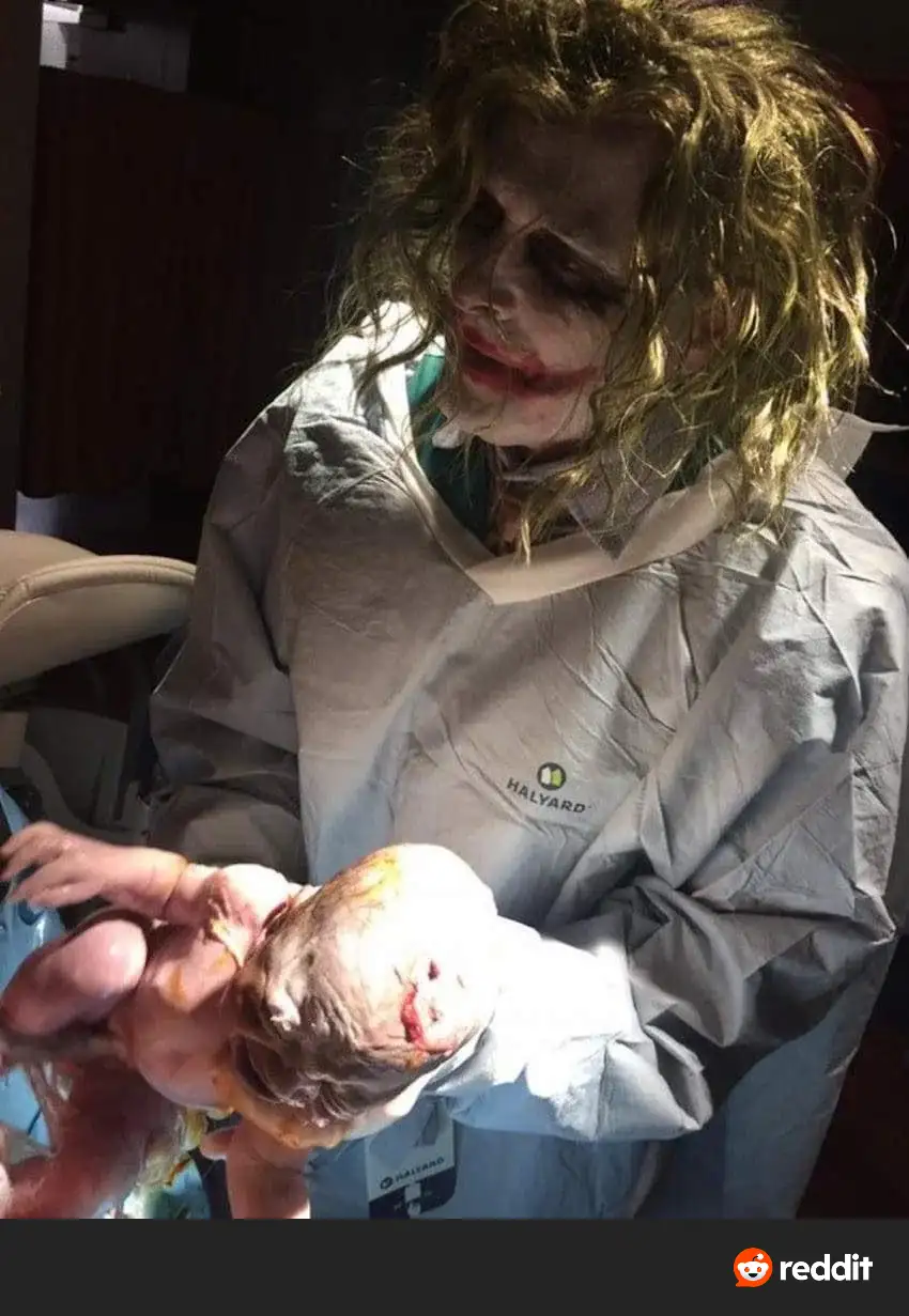 A person dressed as the Joker, wearing a medical gown with 'HALYARD' branding, holds a newborn baby in a clinical setting…