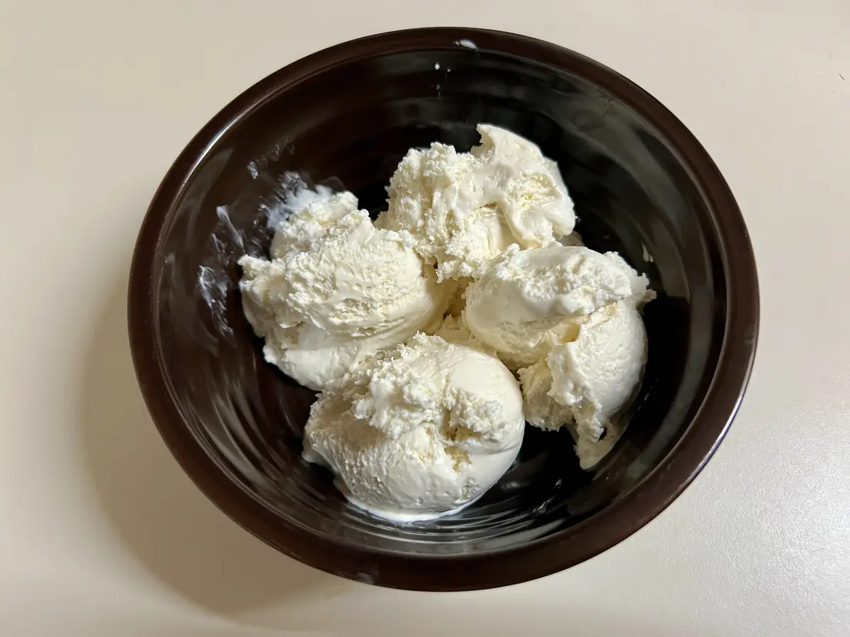 A top-down photo shows several scoops of vanilla ice cream in a dark brown bowl, with some melted drips on the sides. The…
