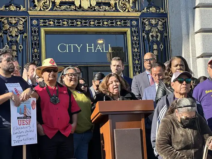 A group of people, including a woman speaking at a podium, stand outside a building with ornate blue and gold doors labeled…