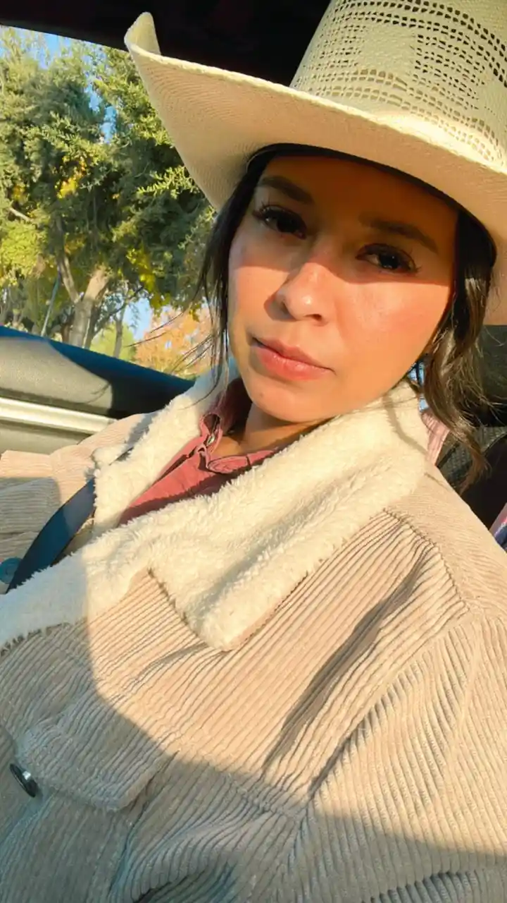 A woman wearing a light-colored cowboy hat and a beige corduroy jacket with a white fleece collar sits in a car, looking at…