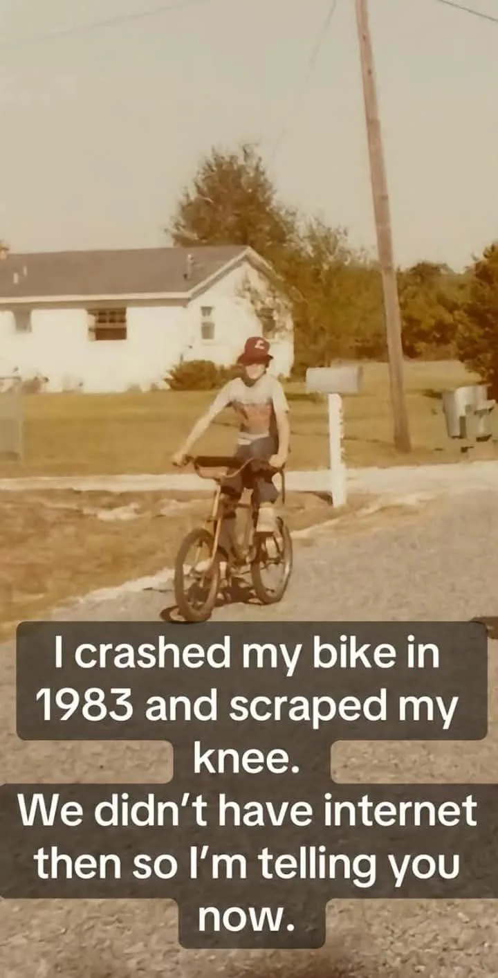 A sepia-toned, slightly blurry photo of a young boy riding a bicycle on a residential street, wearing a red cap and a…
