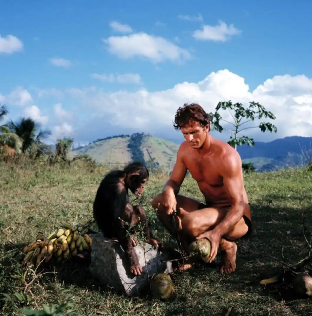 A shirtless, muscular man crouches outdoors in a grassy field under a blue sky with clouds, holding a coconut and a knife…