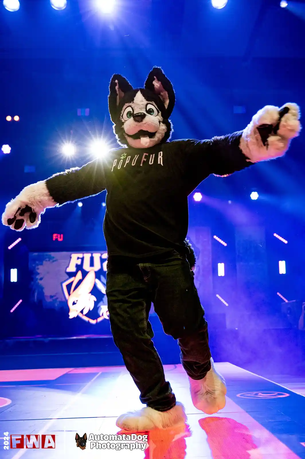 A person in a black-and-white husky fursuit stands on a brightly lit stage with arms outstretched, wearing a black t-shirt…