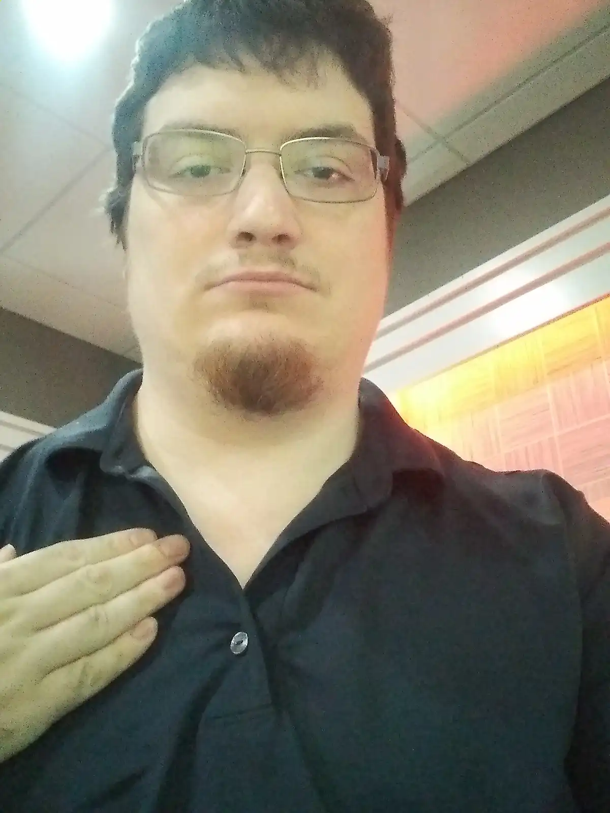 A man with glasses and a goatee, wearing a dark collared shirt, places his hand over his heart while looking directly at the…
