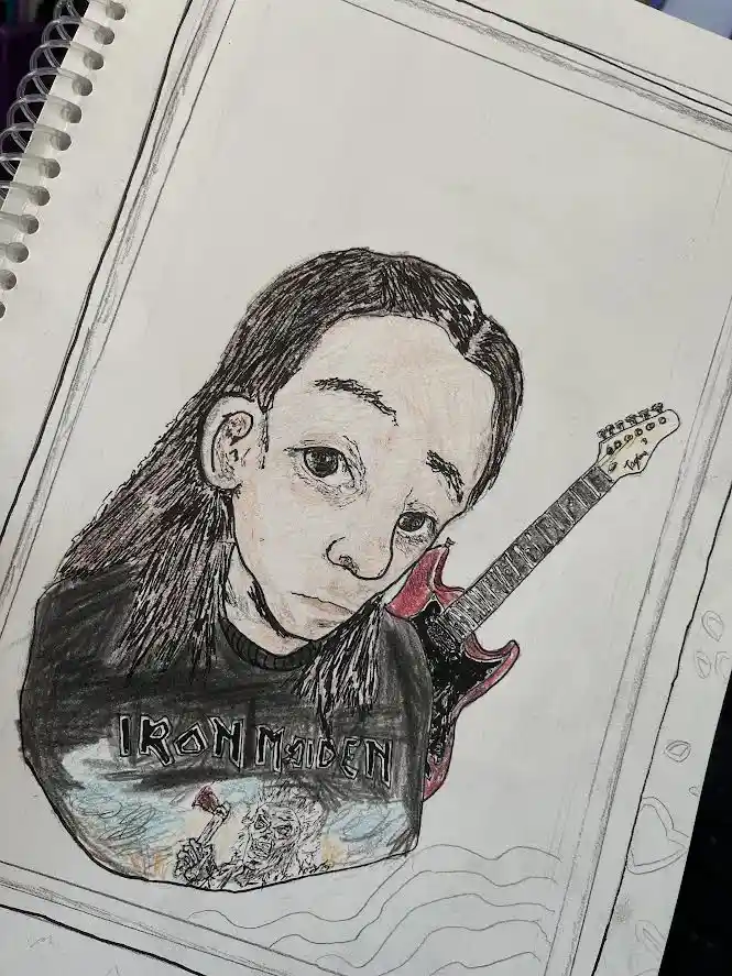 A hand-drawn sketch in a spiral-bound notebook shows a person with long hair wearing a black t-shirt with the 'Iron Maiden'…