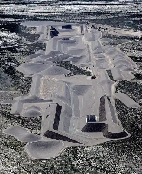 An aerial photograph shows a vast, complex landscape of dirt mounds, berms, and winding tracks carved into a desert-like…