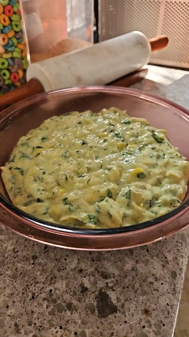 A close-up photo shows a thick, creamy, greenish-yellow mixture with chopped green herbs in a pink glass bowl, sitting on a…