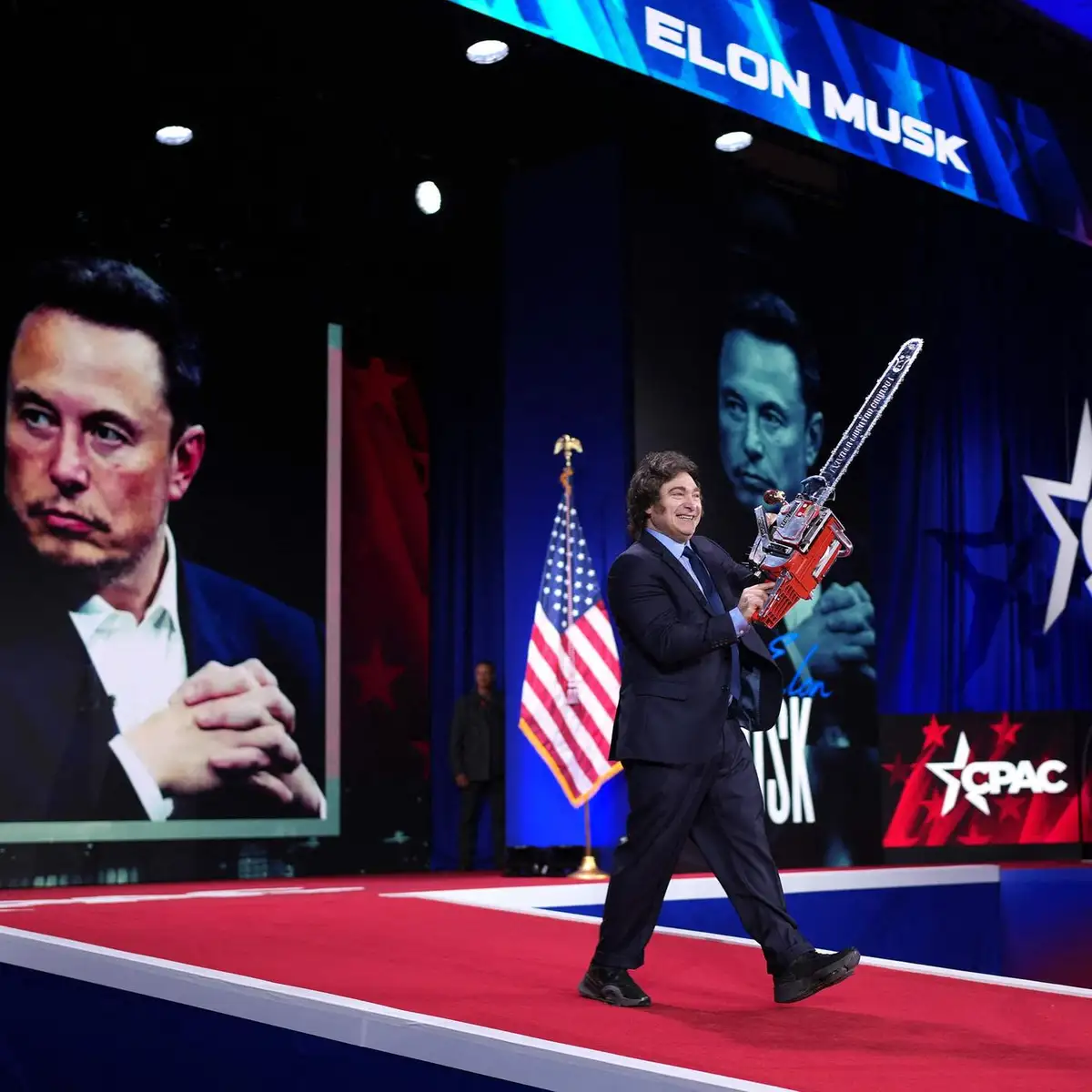 A man in a suit walks across a red carpet stage holding a chainsaw, smiling broadly. Behind him, large screens display a…