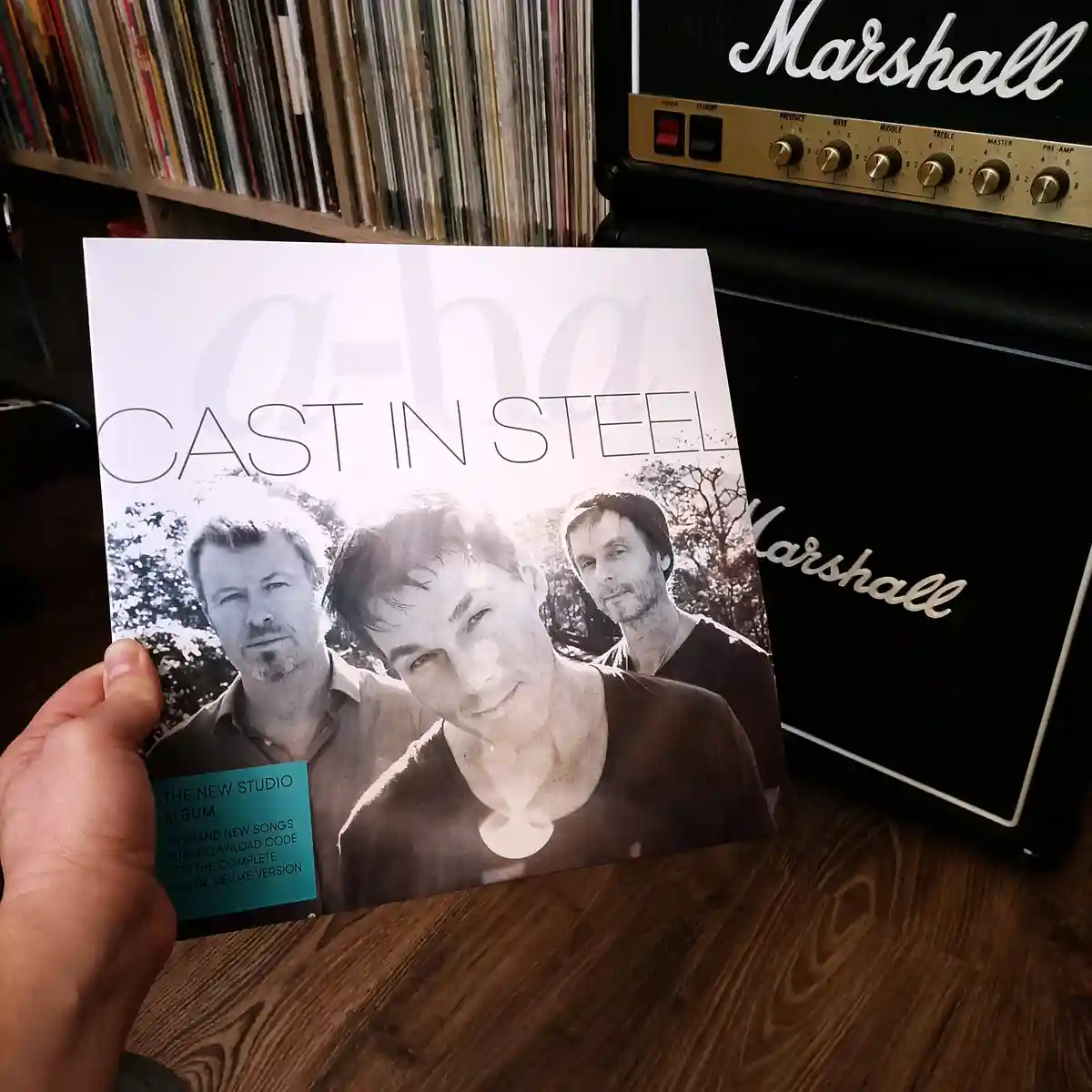 A hand holds up the album cover for a-ha's 'Cast in Steel', featuring a black-and-white photo of the three band members. In…