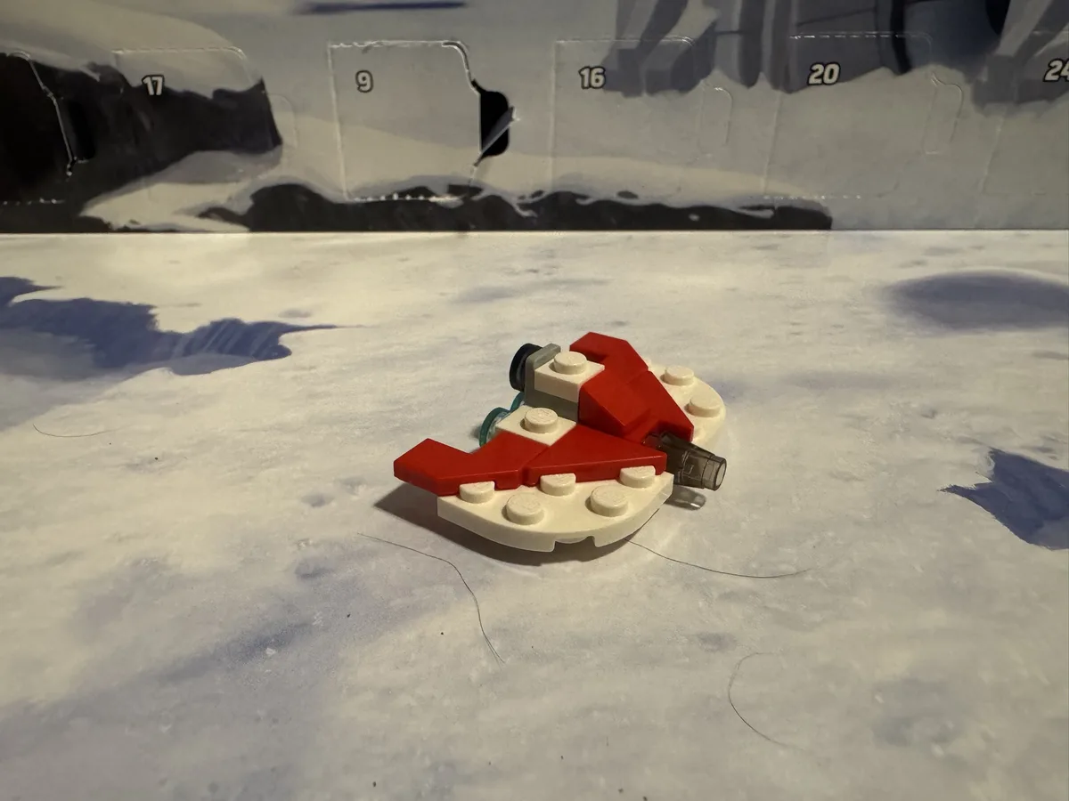 A small, crudely assembled LEGO spaceship model, primarily red and white, sits on a snowy surface in front of a partially…