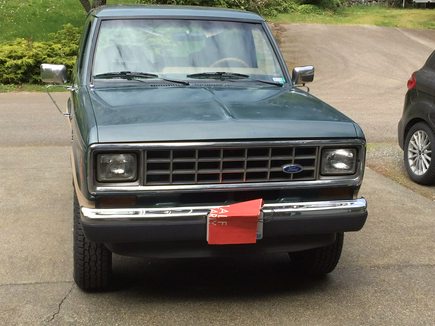 a front-facing photo of a dark green, older model ford truck parked on a paved driveway. The truck has a chrome grille and…