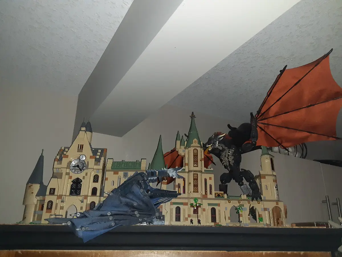 A LEGO model of Hogwarts Castle is shown with two large, toy dragons—one blue and one black with orange wings—positioned as…