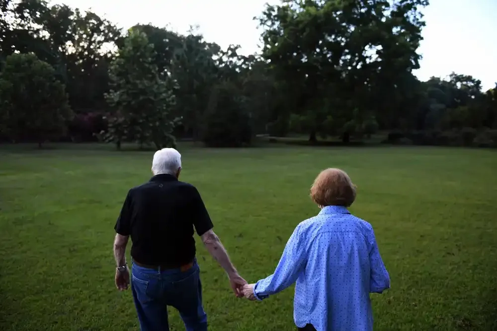 An elderly couple walks hand-in-hand across a grassy park, seen from behind, with trees in the background. The man wears a…