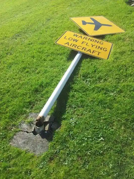A yellow warning sign with a black airplane symbol and the text 'WARNING LOW FLYING AIRCRAFT' is mounted on a white pole…