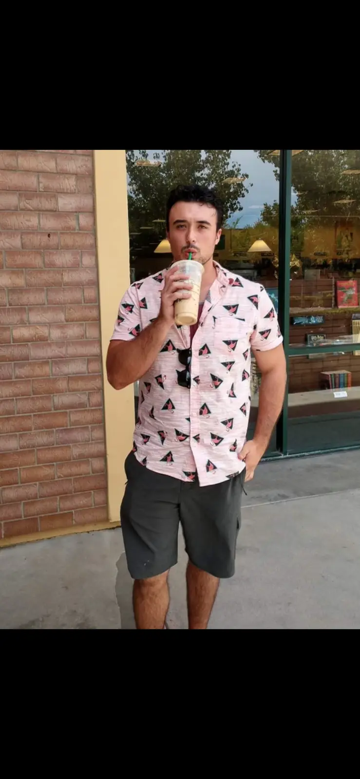 A man in a pink flamingo-patterned shirt and dark shorts stands outside a storefront, sipping a light-colored beverage from…