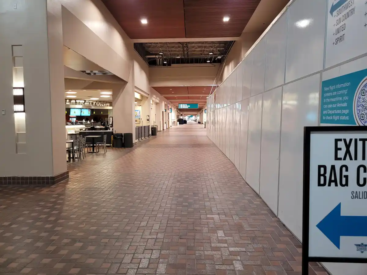 An empty airport corridor with a 'LATRATTORIA' food stall on the left and a long wall of white construction barriers on the…