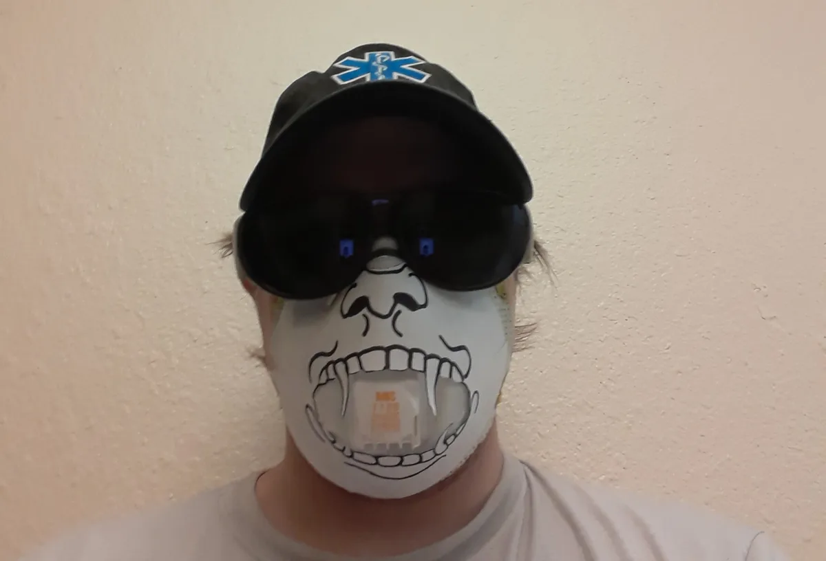 A person wearing a black baseball cap with a blue medical symbol, large black sunglasses, and a white face mask drawn with a…