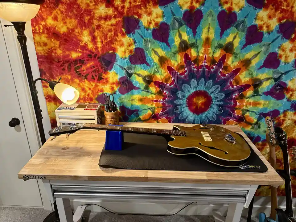 A sunburst electric guitar rests on a black mat atop a light wood Husky workbench, propped up by a blue stand. Behind it, a…
