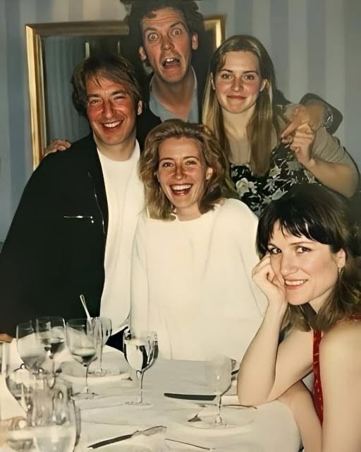 A group of five people are posing together at a dinner table, with wine glasses and cutlery visible. The person in the back…