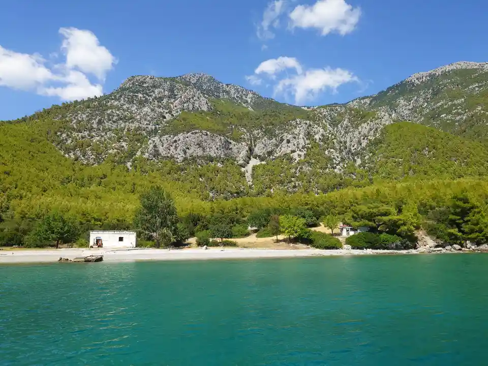 A scenic view of a calm turquoise sea meeting a pebbled shore with small white buildings and lush green trees, backed by…