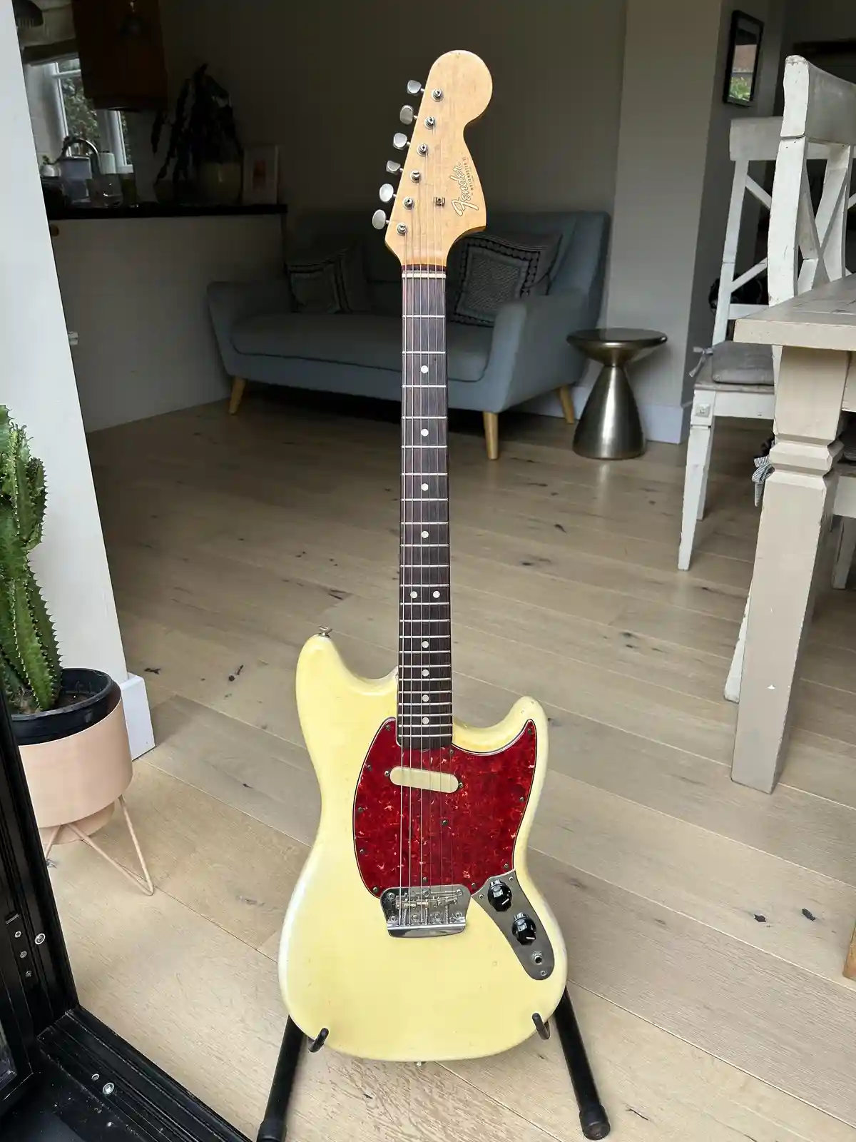 A vintage-style Fender electric guitar with a cream body and red pickguard stands upright on a stand in a modern home with…