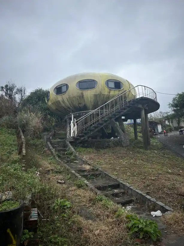 A weathered, yellow, egg-shaped structure resembling a spaceship sits atop a hill, accessed by a metal staircase and stone…