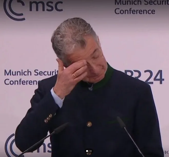 A man in a dark jacket with a green collar stands at a podium with microphones, visibly distressed as he rubs his eye with…