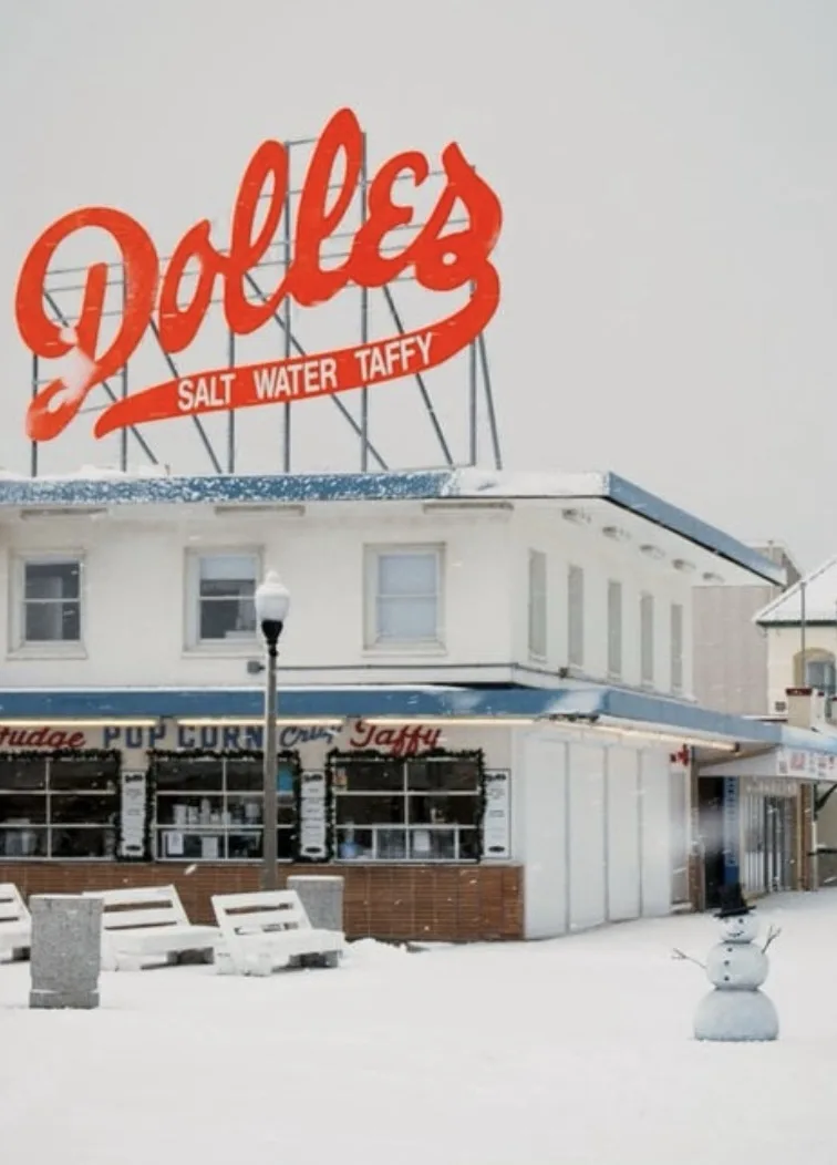 A snowy scene shows Dolles Salt Water Taffy store with its large red sign, snow-covered benches, and a small snowman in the…