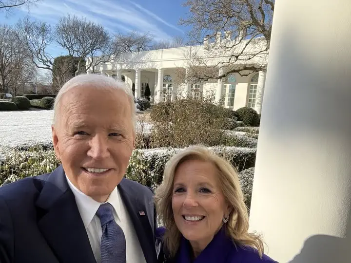 A selfie of Joe Biden and Jill Biden smiling in front of the White House, with snow on the ground and bare trees in the…