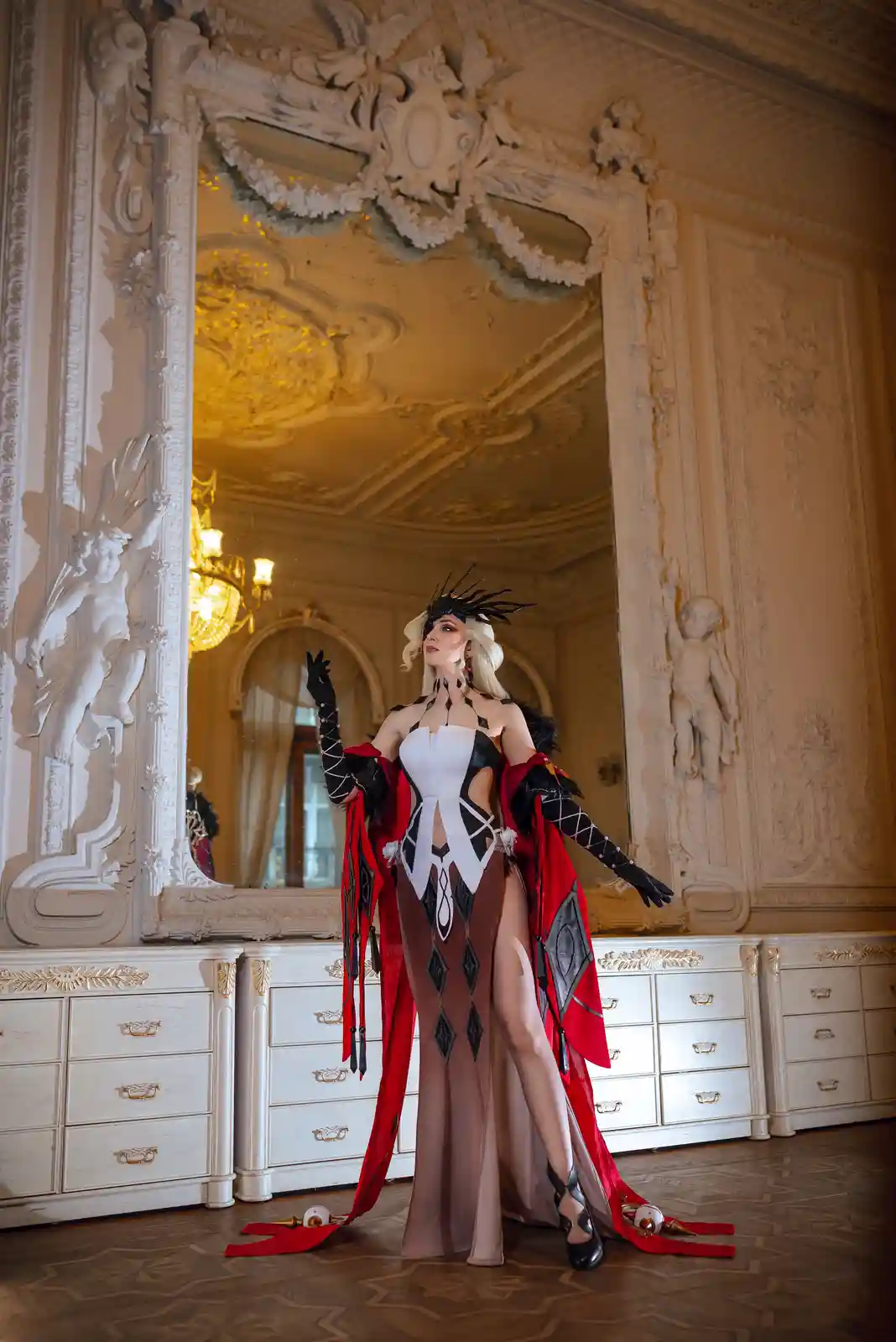 A cosplayer in an elaborate black, white, and red costume with a dramatic headdress and long gloves poses in a grand, ornate…